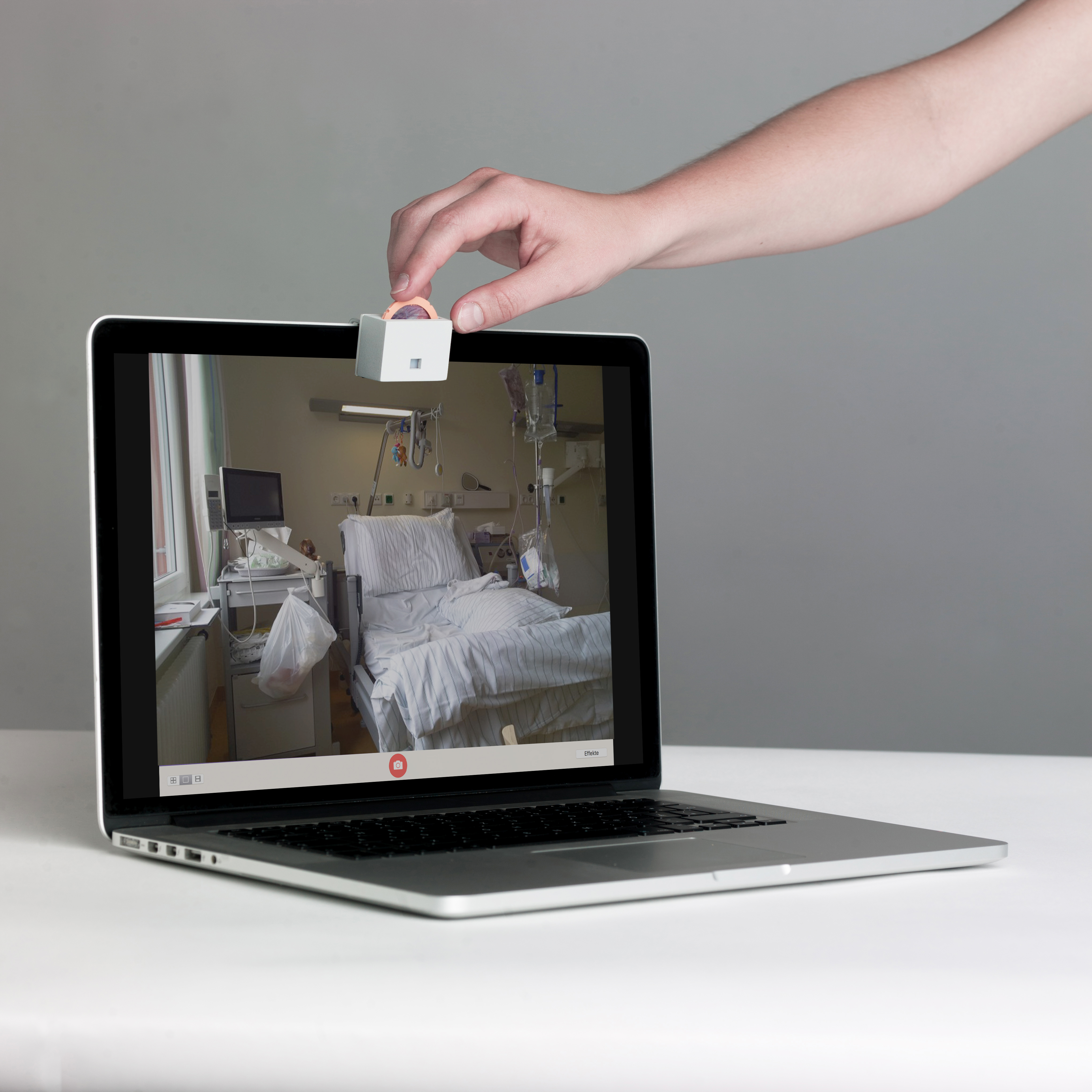 Photo of »Object A – Un-hacking Webcams« of the work »Accessories for the Paranoid«, laptop with a small device with a dial in front of the webcam, on which a hand turns. On the screen you can see a hospital bed room.