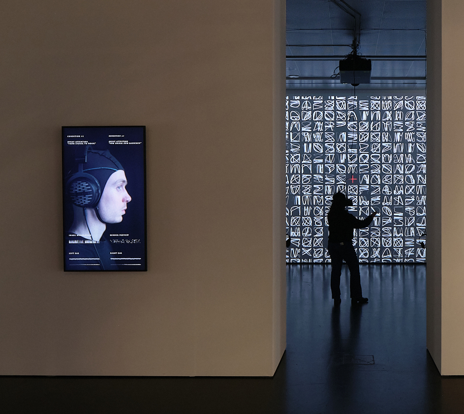 »AUTOPOIESIS« von ATELIER-E. Es sind zwei Räume, verbunden durch einen Durchgang zu sehen. An der Wand des vorderen Raumes befindet sich ein Bildschirm, auf dem eine Person mit einer Haube abgebildet ist. Im zweiten Raum steht eine Person mit VR-Brille.
