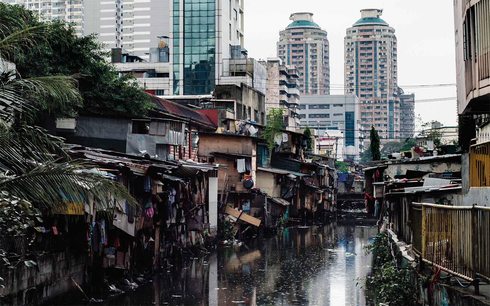 The picture shows dilapidated huts on a dirty river in front of modern high-rise facades. 
