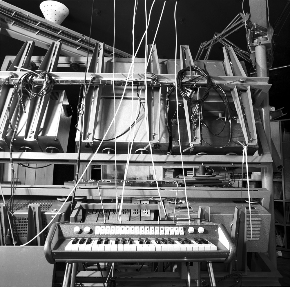 A black and white picture shows an electric piano in front of cables and computers. 
