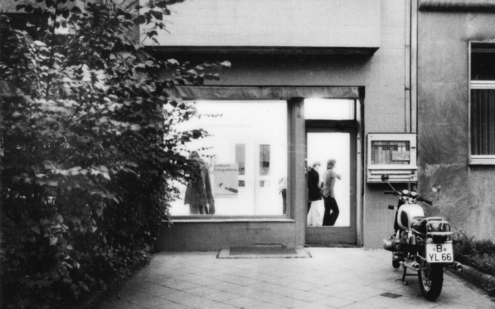 Schwarz-weiß Foto zeigt die Straßenansicht eines Galerieraum. Davor steht ein Motorrad.