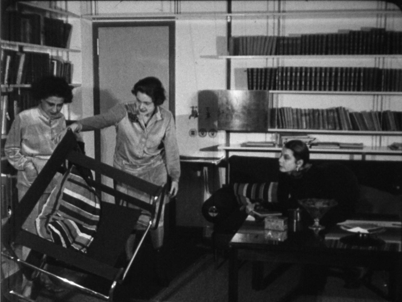 Filmstill aus »Wie wohnen wir gesund und wirtschaftlich?« von 1926 in schwarz-weiß. Darauf sind drei Frauen zu sehen, die die Möbel in einem Wohnzimmer begutachten.