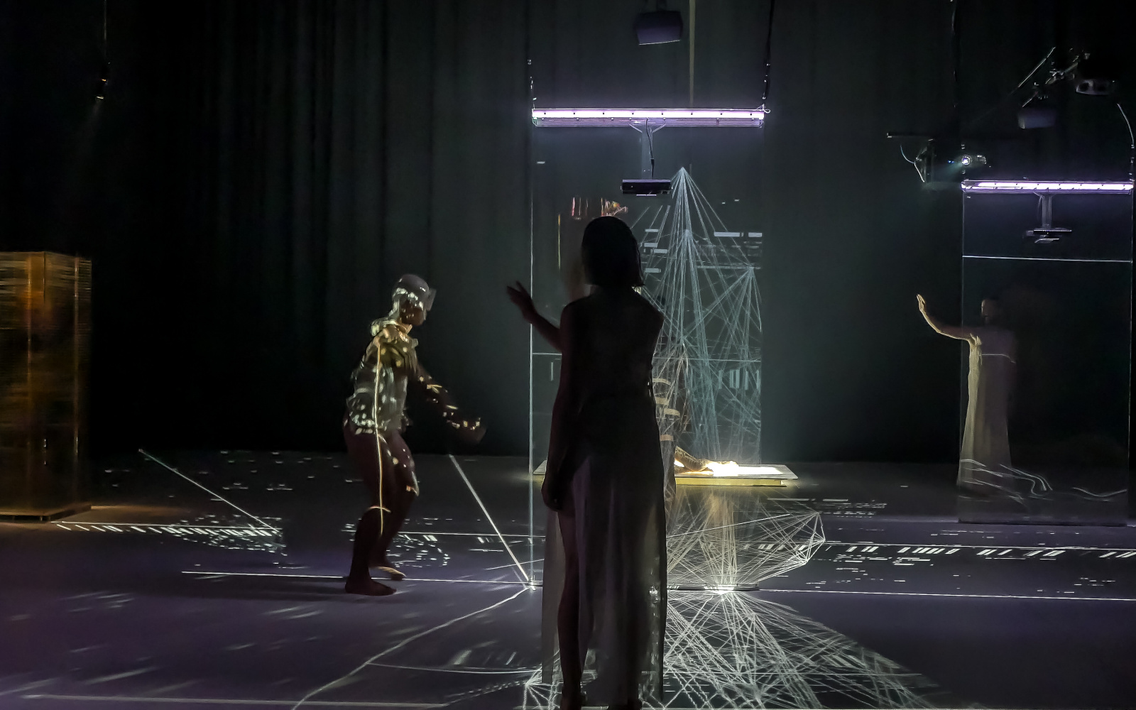 Three dancers stand on the stage, it is dark. In the middle of the stage is an installation of laser light. On the body of the left dancer is a projected play of light.