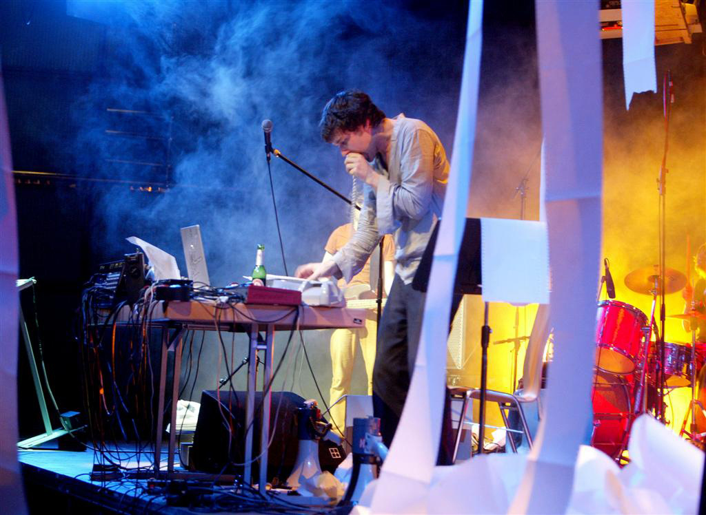 Jamie Lidell performs on stage. From the printers hanging above the stage printers fan-fold paper drops.