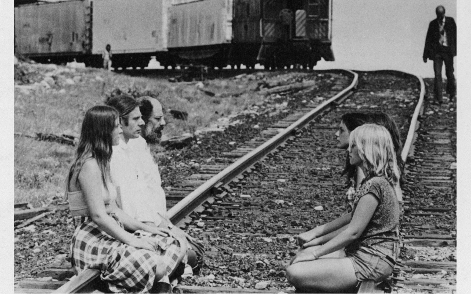 Auf einem Bahngleis sitzen sich Menschen gegenüber. Von hinten naht ein Zug.