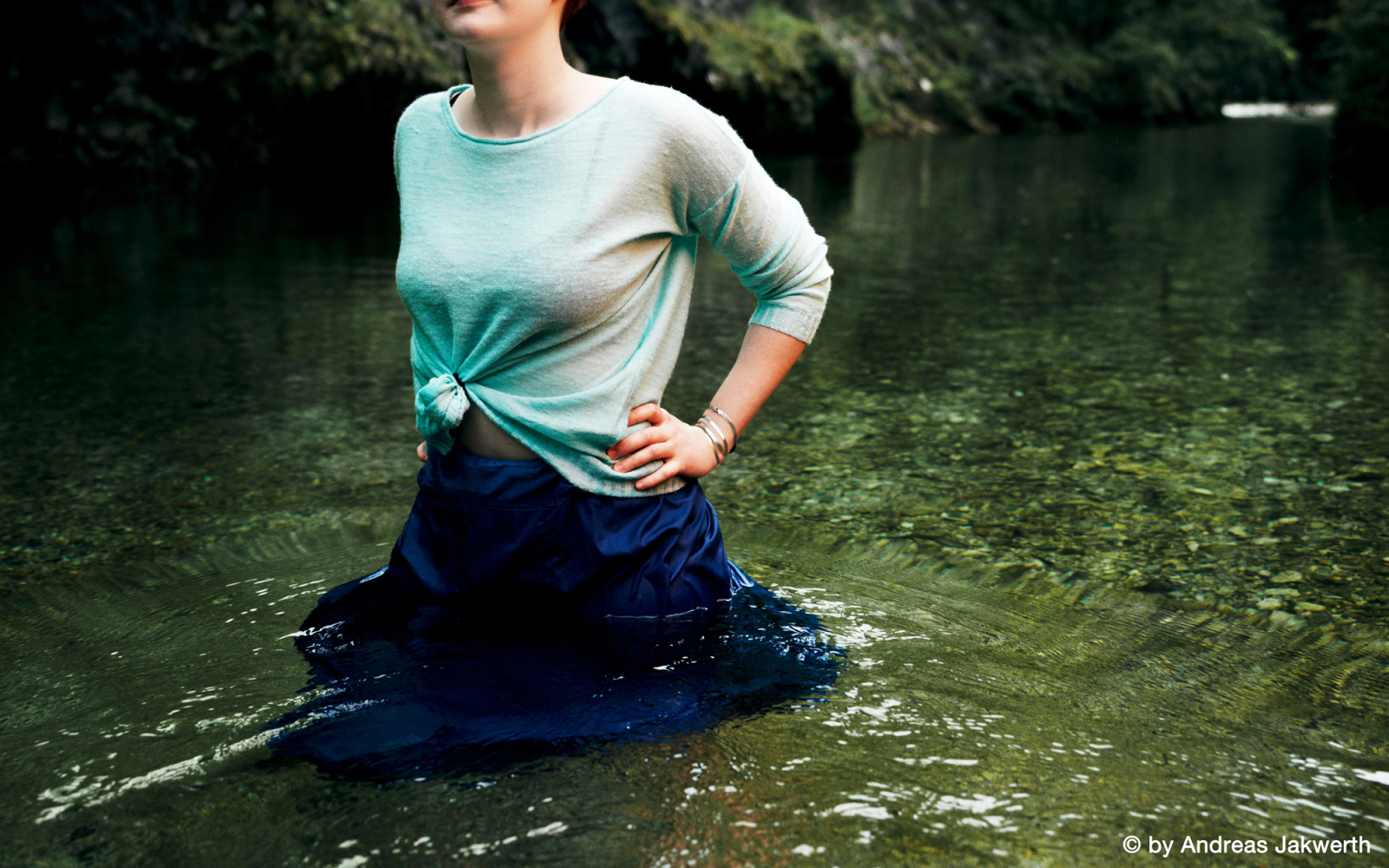 A woman is standing in the water. She is wearing a blue skirt and a mint green sweater. Your hair is red. Water and landscape seem to green.
