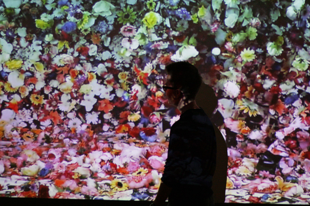 A woman stands in front of a canvas on which many colorful flowers are innumerable to see.