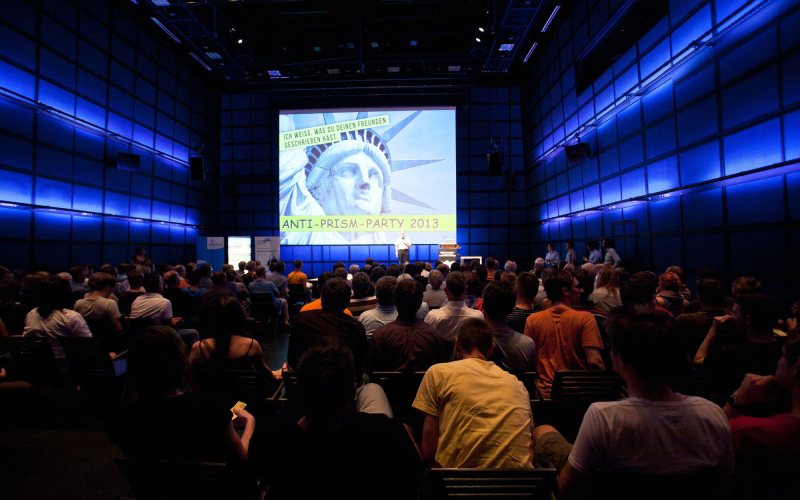 People sitting in front of a stage. On the screen of the head of the Statue of Liberty can be seen