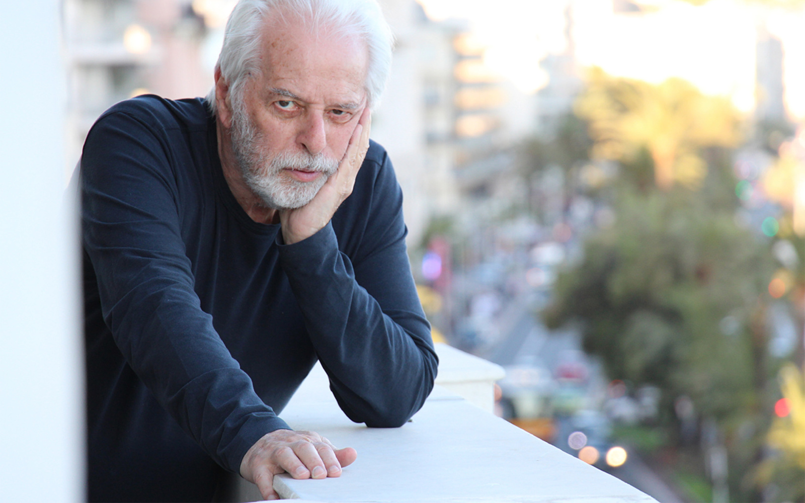 A man leans on a railing. He is wearing a dark sweater. His hair and beard are gray.