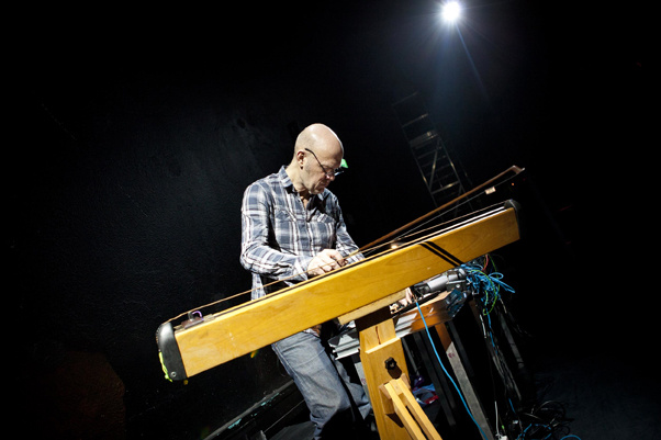 Der Hintergrund ist schwarz. Ein Mann mit Brille, bekleidet mit einem Hemd und Jeans, spielt ein Instrument und bedient dazu ein Mischpult.
