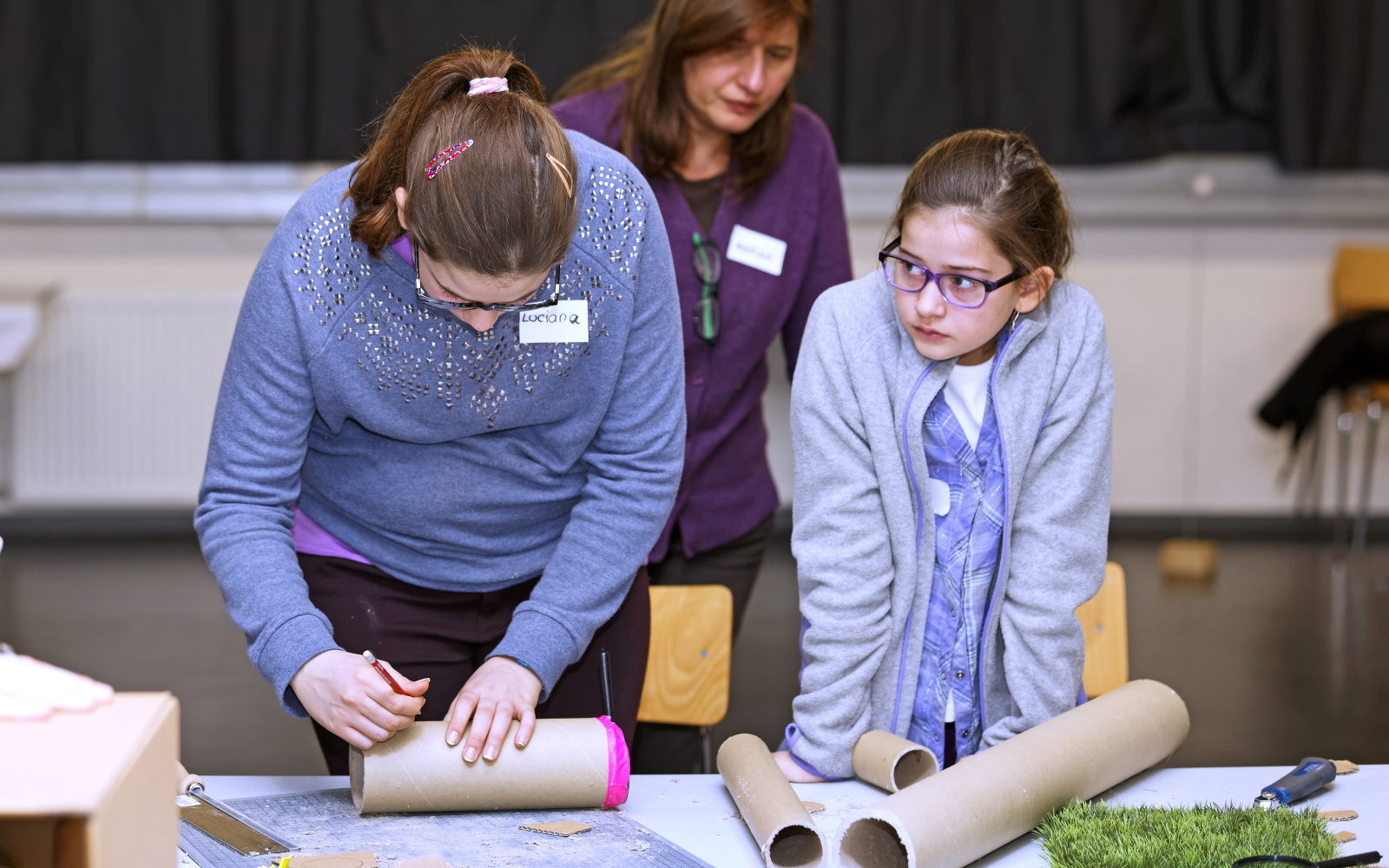 Two girls are standing in front of a table with cardboard-rolls. One of them is marking the roll in front of her with a pencil.