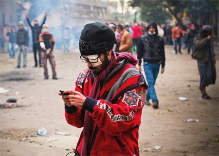 A man in a red jacket and black hat. His left eye is connected. He wears glasses and looks at something he holds in his hands. In the background, a large number of people.