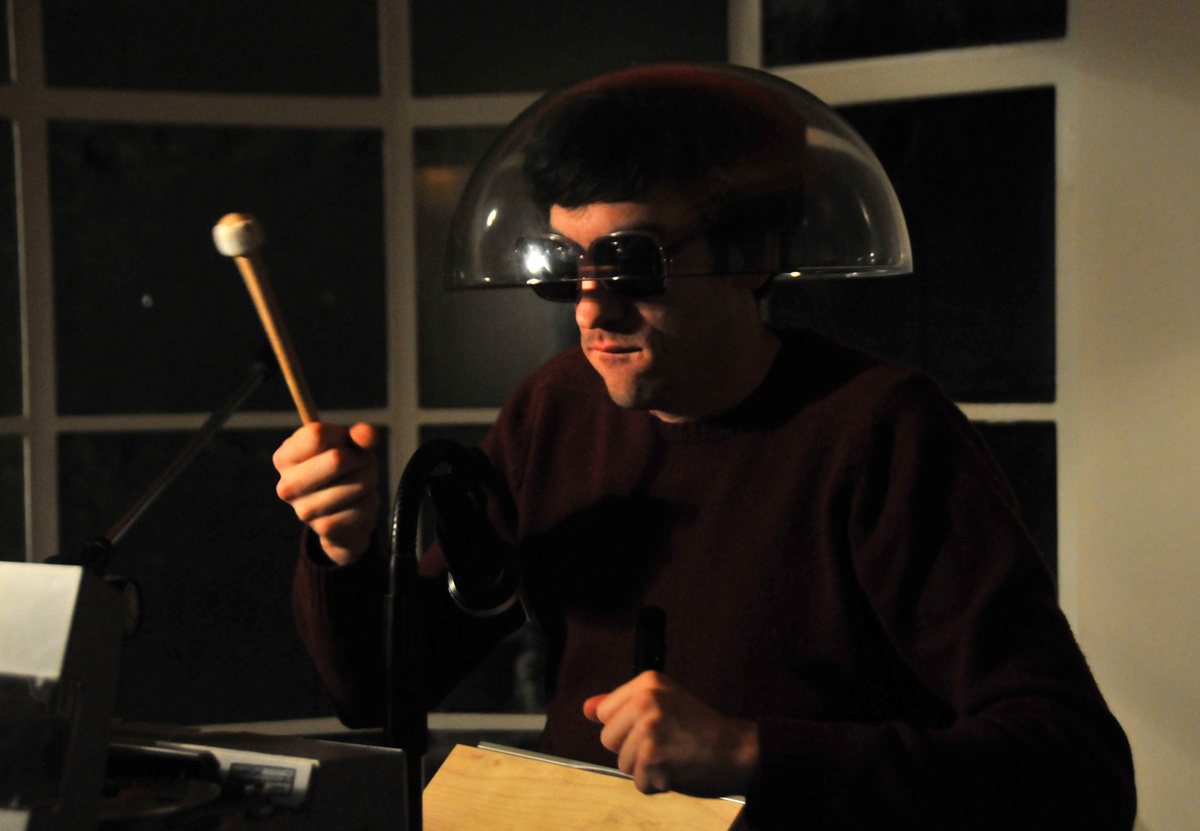 A man wearing sunglasses and transparent lampshade on his head, holding a drumstick in his right hand.
