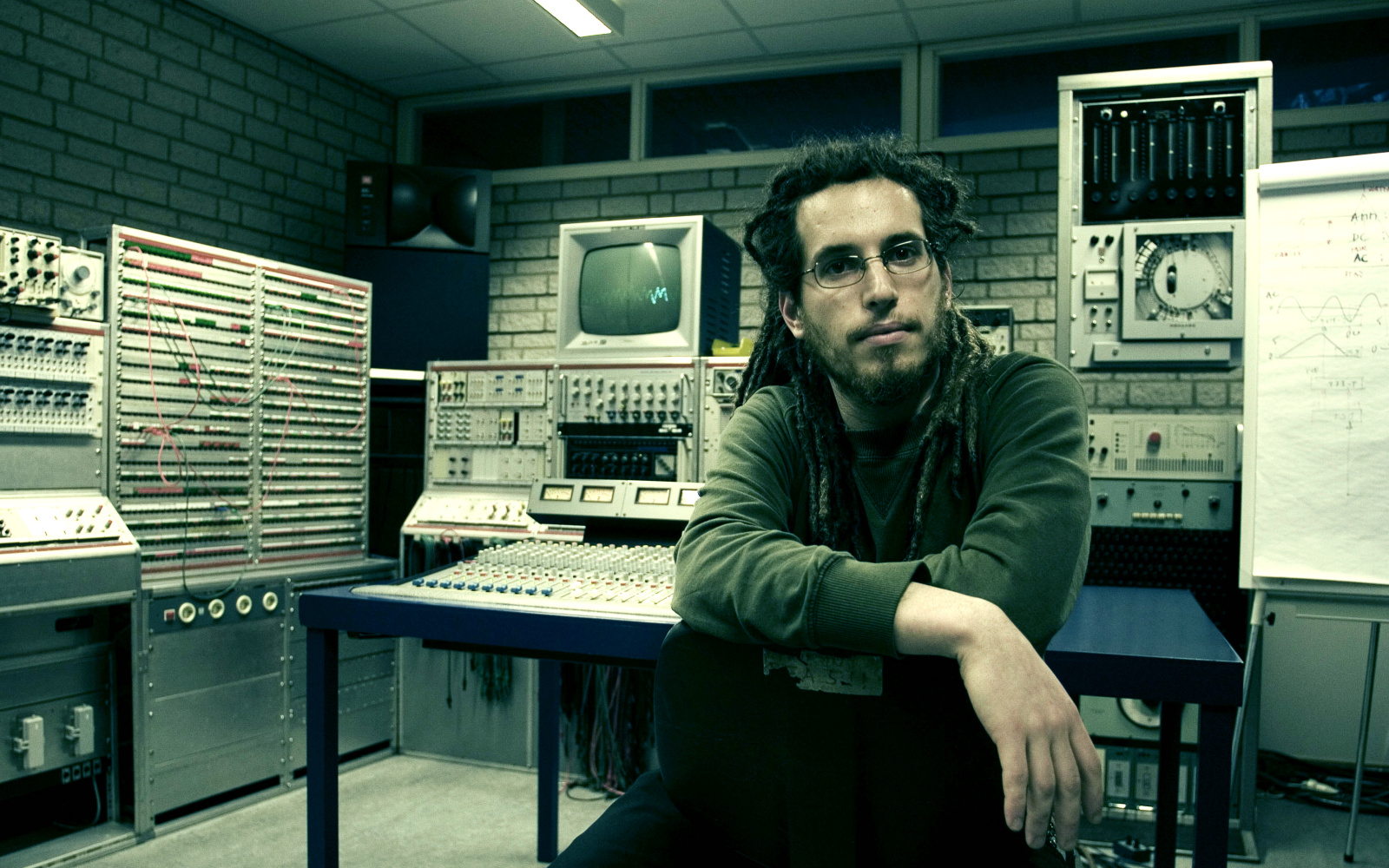 A man with dreadlocks and glasses sitting in front of technical equipment such as PCs, volume controls and other monitors