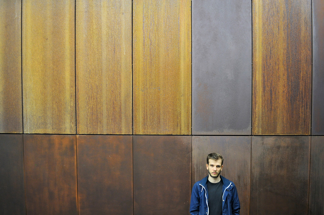 A young man wearing blue sports jacket standing in front of a wooden wall