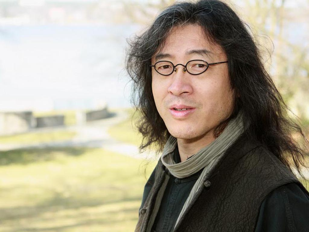 A man with glasses and long hair stands in front of a tree