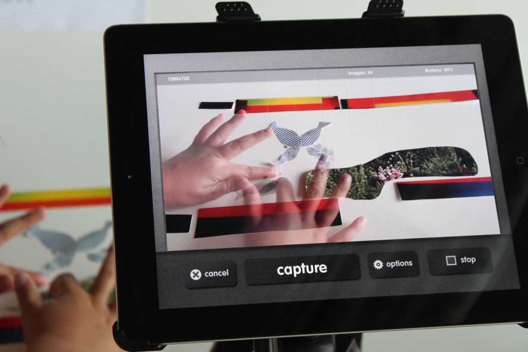 Per iPad werden zwei Kinderhände aufgezeichnet, die an einer Papiercollage arbeiten.