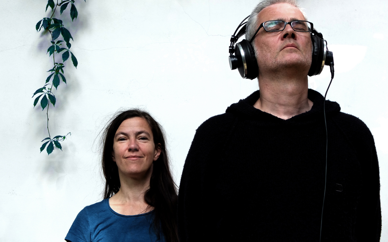 A man with closed eyes and headphones stands right in front of a woman who looks at the camera