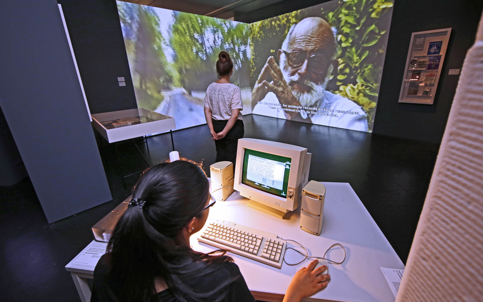 Exhibition view: foreground a girl sitting at a computer, in the background a video projection with Vilém Flusser that fills an entire wall