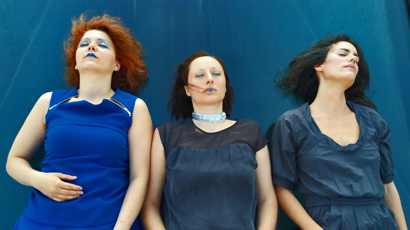 Three women dressed in blue and wearing blue makeup, lying on the blue tarp