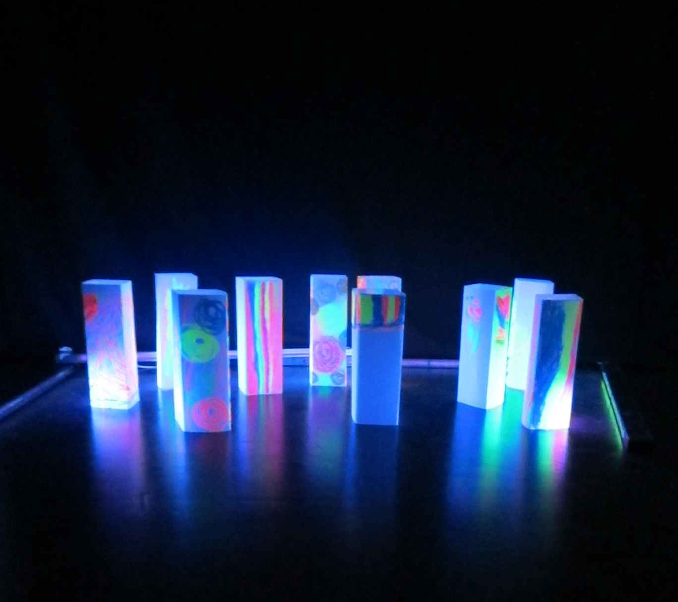 Numerous, with bright colors painted blocks are glowing in the neonlight. They stand in front of a black background and resemble a skyline.