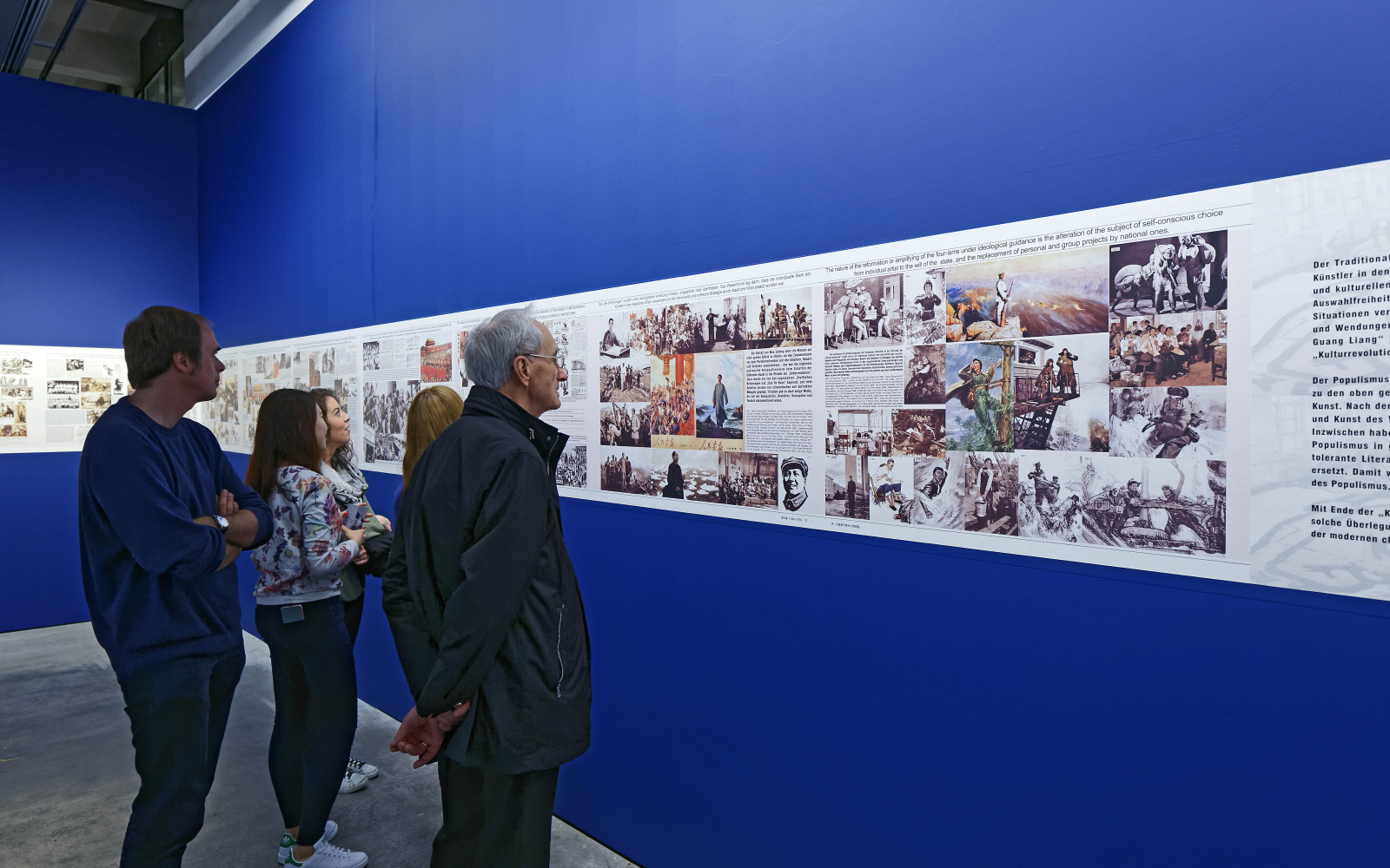 Fünf Menschen betrachten Bilder in einer Ausstellung, die auf blauem Grund hängen