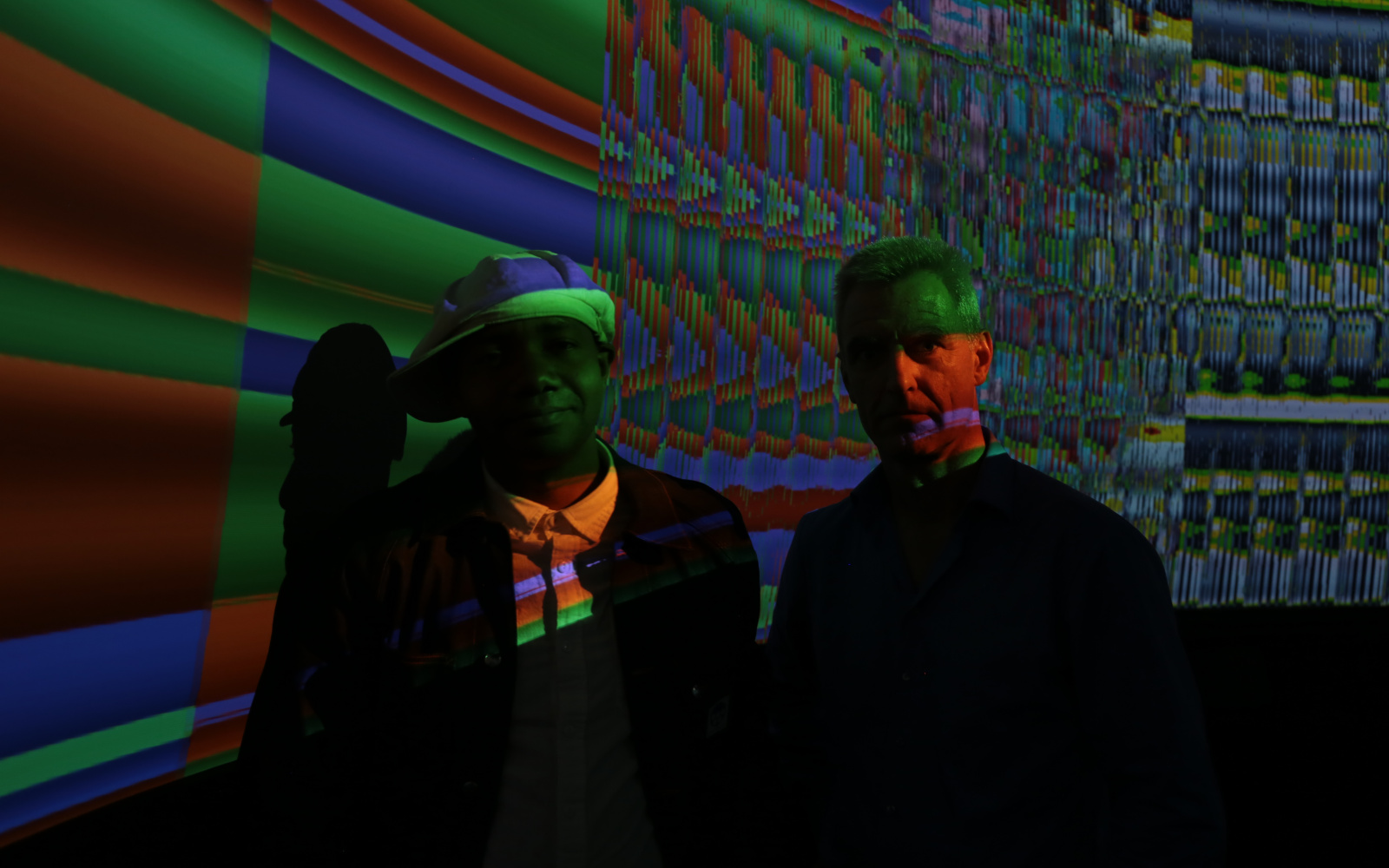 Two men standing in a dark room in front of a screen with colorful colors.