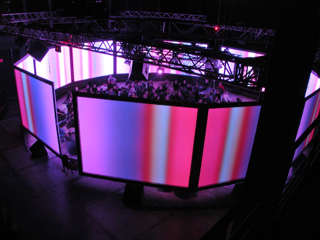Circularly arranged, colorful illuminated screens in the middle of which a group of people is sitting.