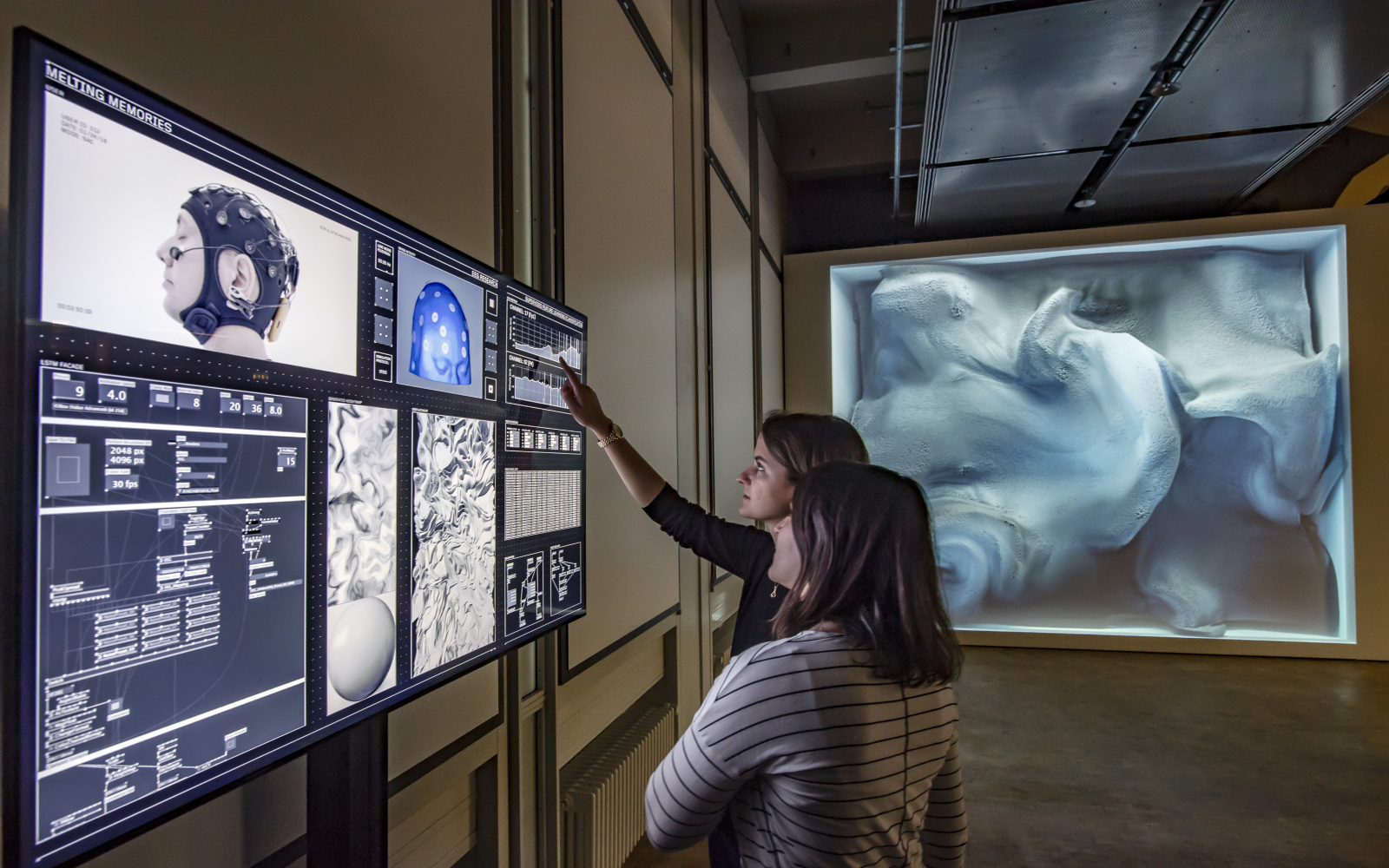Zwei Frauen stehen vor einem Bildschirm, der einen Kopf, der an ein Elektroenzephalogramms (EEG) angeschlossen ist, und die daraus resultierenden Daten zeigt.