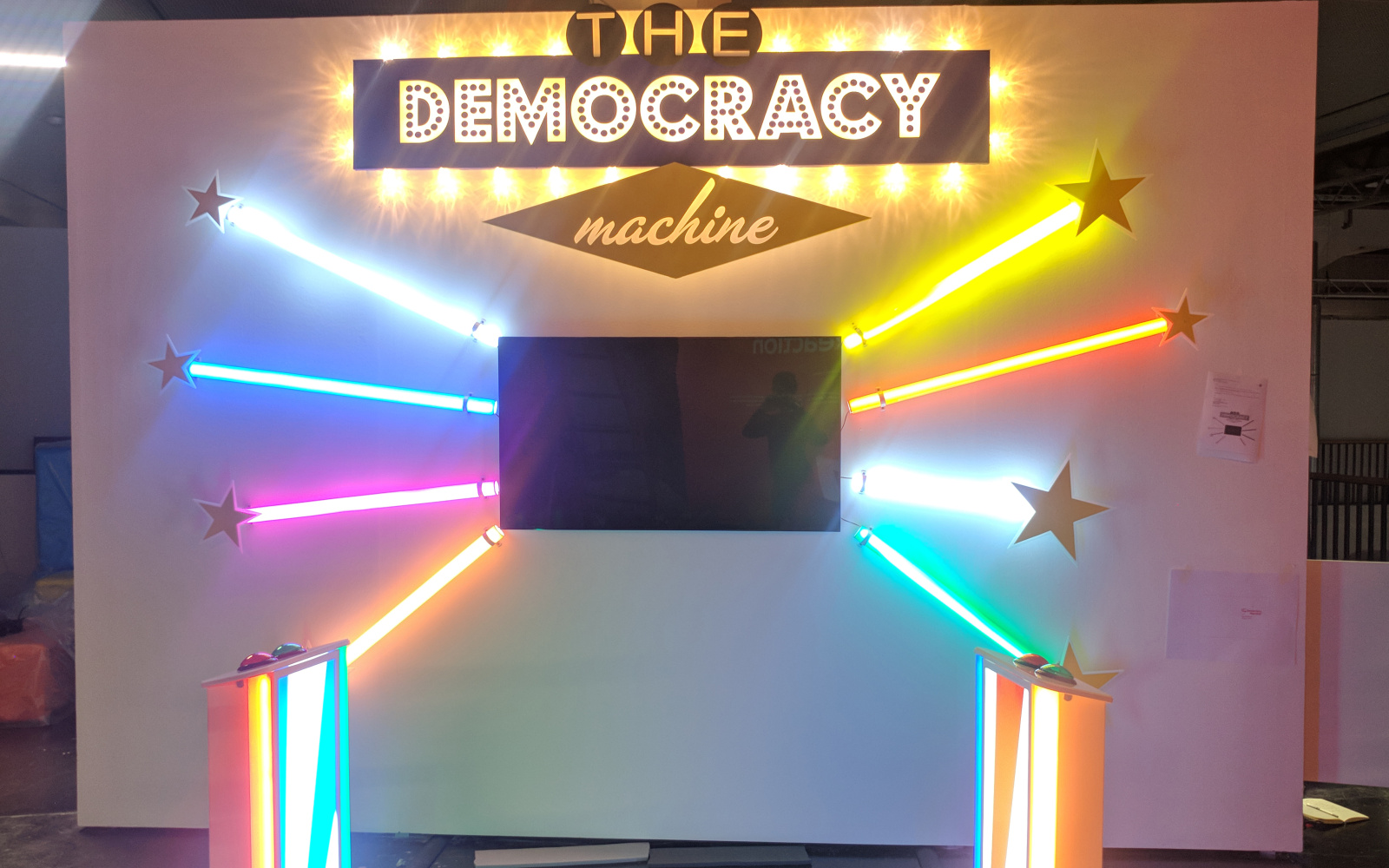 Frontalansicht der The Democracy Machine!. In der Mitte einer Wand ist ein Fernseher installiert darum herum sind je drei bunte Farbröhren wie Sonnenstrahlen angeordnet. Davor stehen zwei Podeste mit Knöpfen.