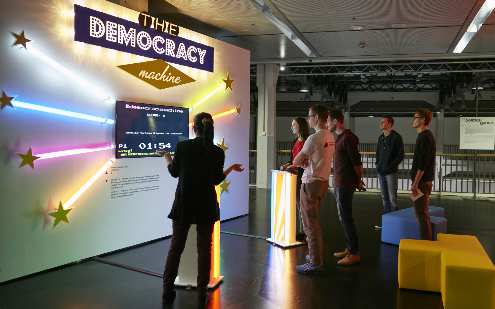 An einer weisen Wand sind ein Bildschirm, bunte Leuchtstäbe und die Leuchtschrift The Democracy Machine angebracht. Zwei Besucherinnen stehen vor den Bedienelementen der performativen Skulptur von Adam Scarborough. Weitere Besucher  stehen etwas abseits.