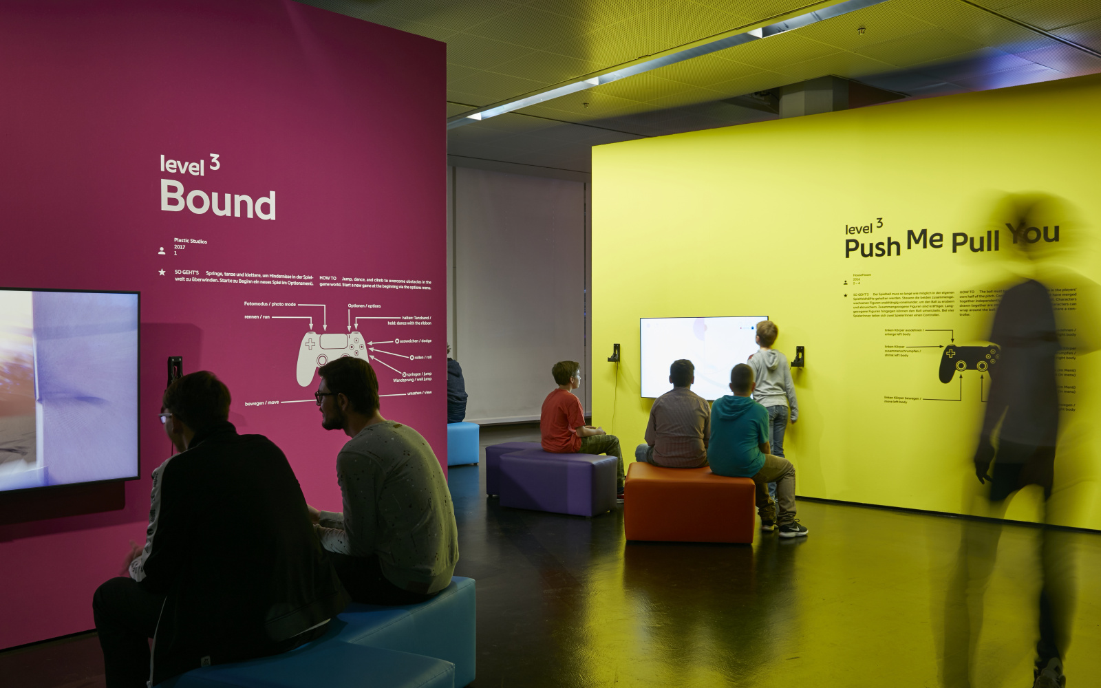 To the left in the foreground, two visitors sit on a bench in front of a screen attached to a collored wall. On the right in the background there are several young visitors sitting in front of a yellow wall with a screen.