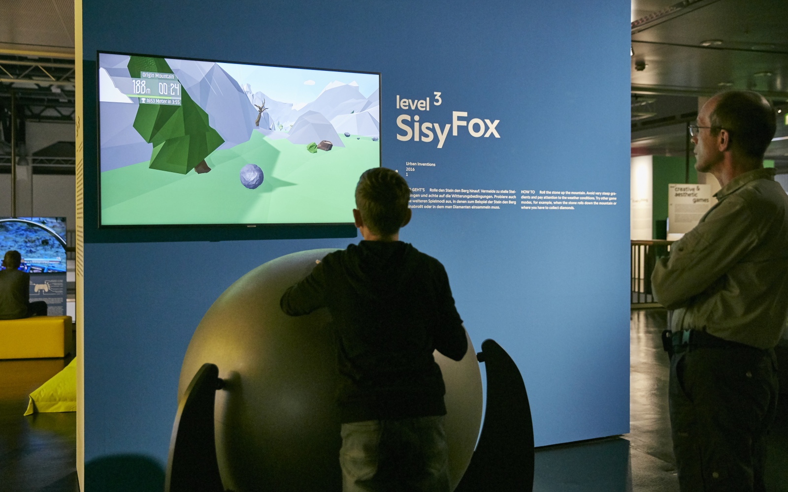 A young visitor in front of a game console consisting of a large ball on a rack. Behind a screen on a blue wall. Another visitor is standing next to it. 