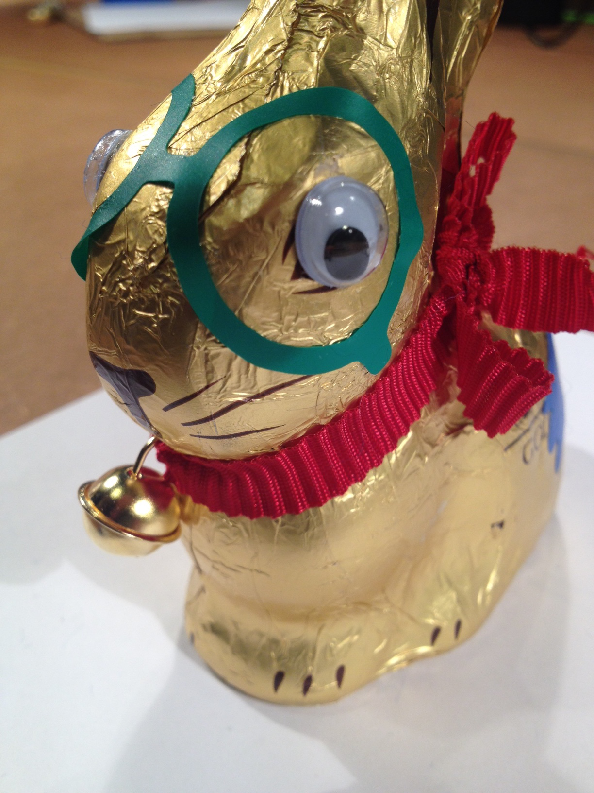 A golden chocolate Easter bunny looks into the camera with its wobbly eyes glued on. Beside the eyes a green sticker similar to glasses is glued on the packaging of the Easter bunny.
