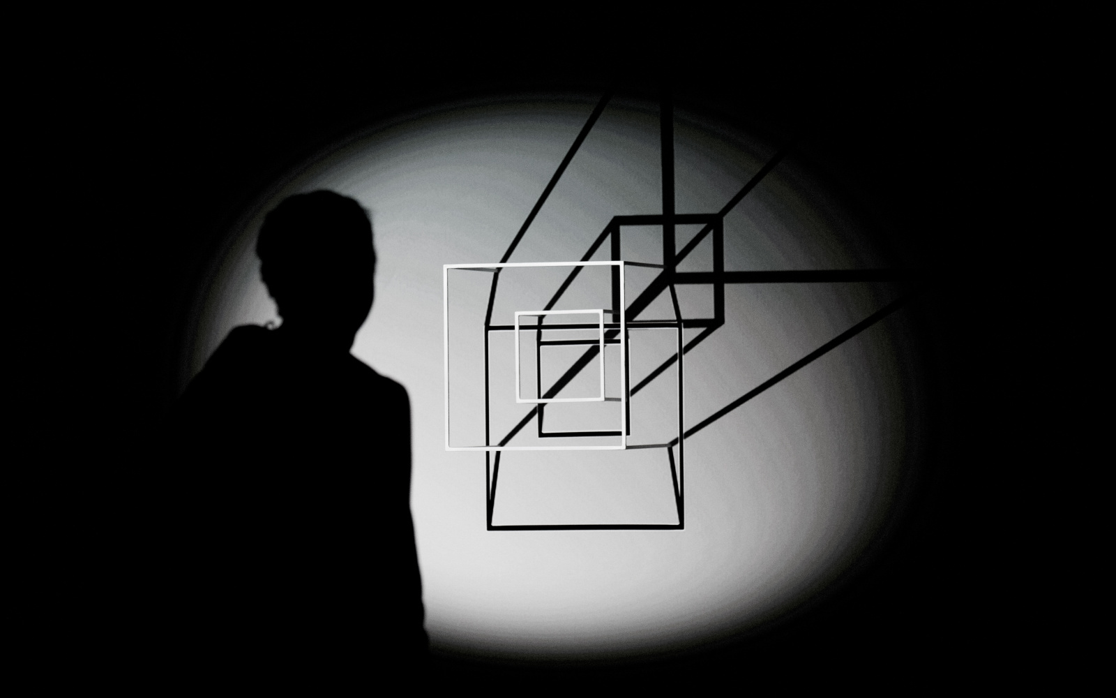 In einem Lichtkegel sieht man den Schatten einer Person und zwei weiße Vierecke, die ebenfalls einen schwarzen Schatten an die Wand werfen