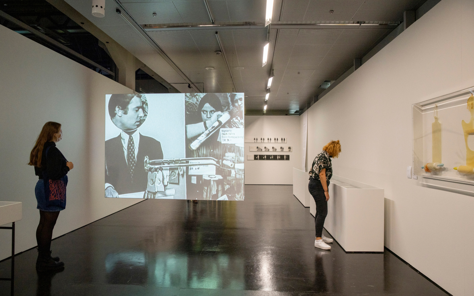 Two young women in the exhibition space. One is standing on the left wall and the other is looking at the right wall in showcases. A screen is stretched between them, on which a film clip with people can be seen.