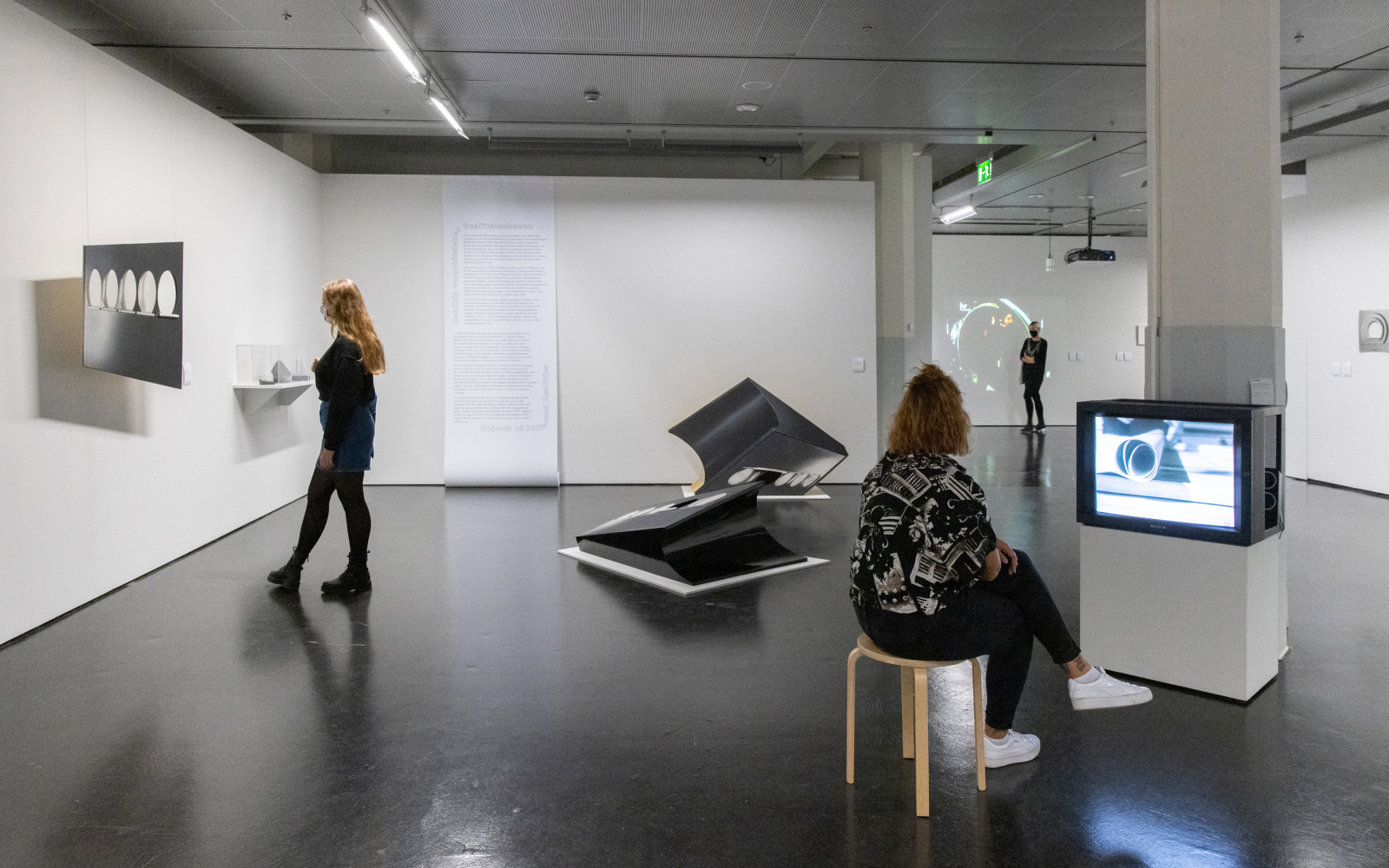 Three women in the exhibition. The woman on the left is looking at a sculpture that is hung on the wall, the woman on the right is watching a film on a TV, in the middle are abstract figures, on the back wall is a wallpaper roll with text on it.