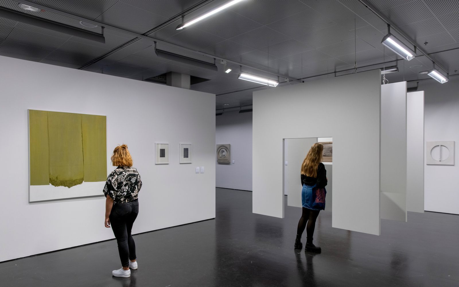 Zwei Frauen im Ausstellungsraum. Links betrachtet eine Frau ein monochromes Gemälde an der Wand. Mittig steht eine Frau vor einer Hängekonstruktion aus drei hintereinanderfolgende Flächen, mit einem Durchgang dazwischen.