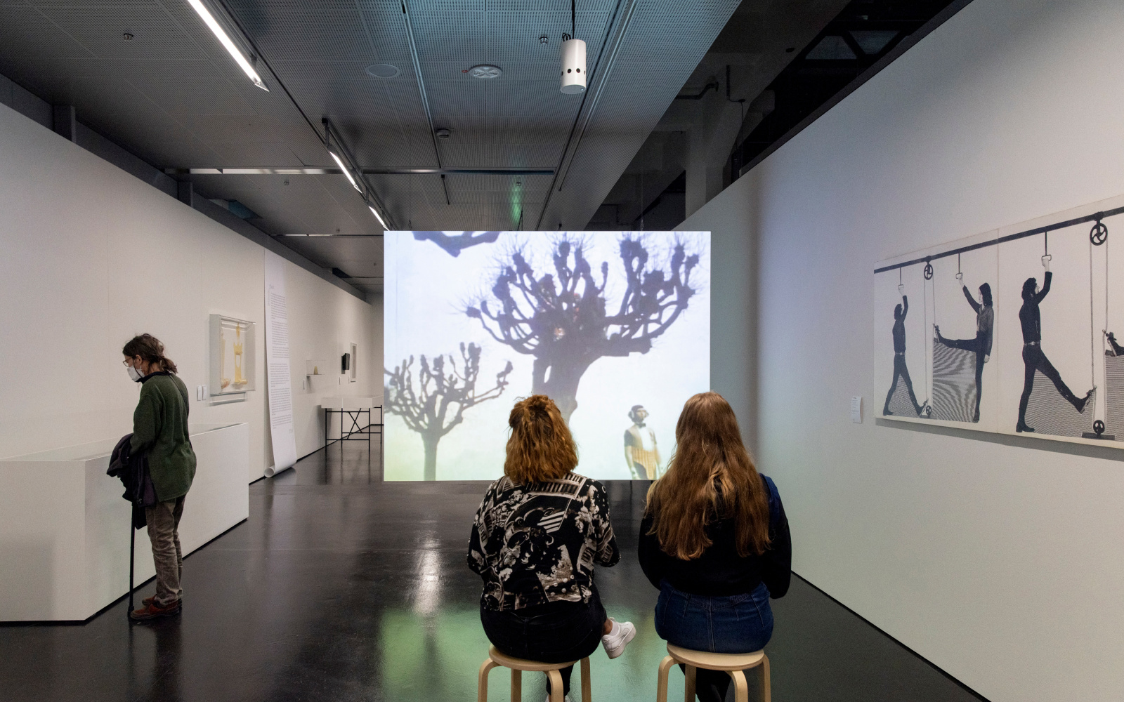 Three young women in the exhibition space. Two sit on stools in front of a film screen on which bare trees can be seen. An abstract drawing hangs on the wall to the right. On the left wall are boxes into which the third woman looks.