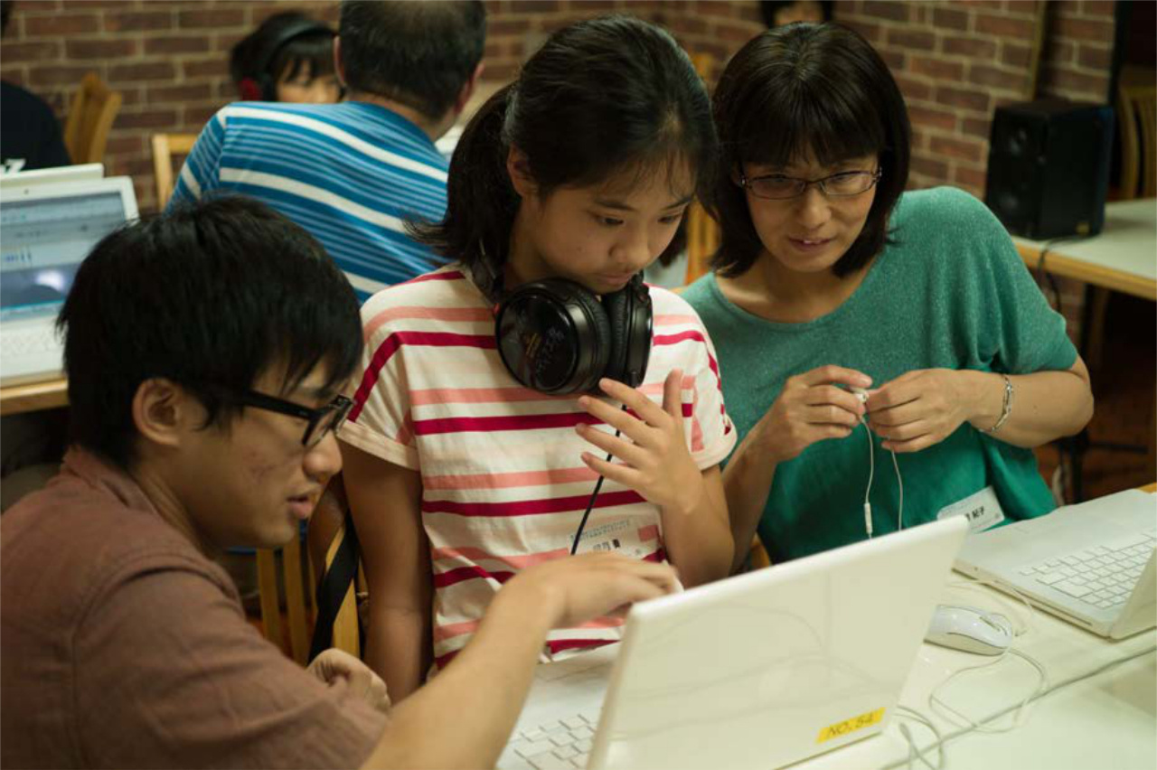 Daughter and mother sit together at a table in front of two laptops. A man kneels next to the table and explains something to them on the computer.