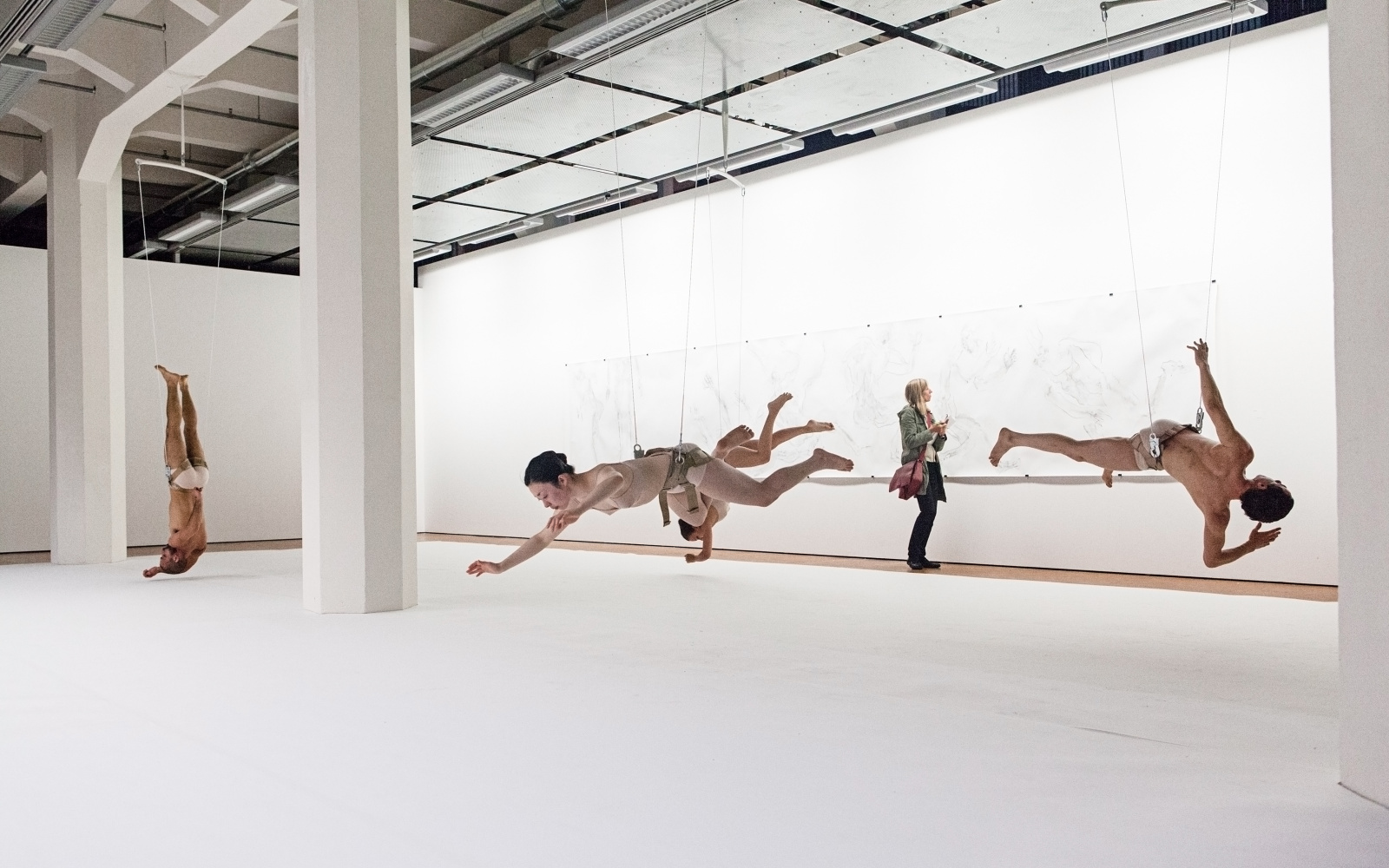 Fünf Tänzer hängen an Drahtseilen kopfüber im Ausstellungsraum.