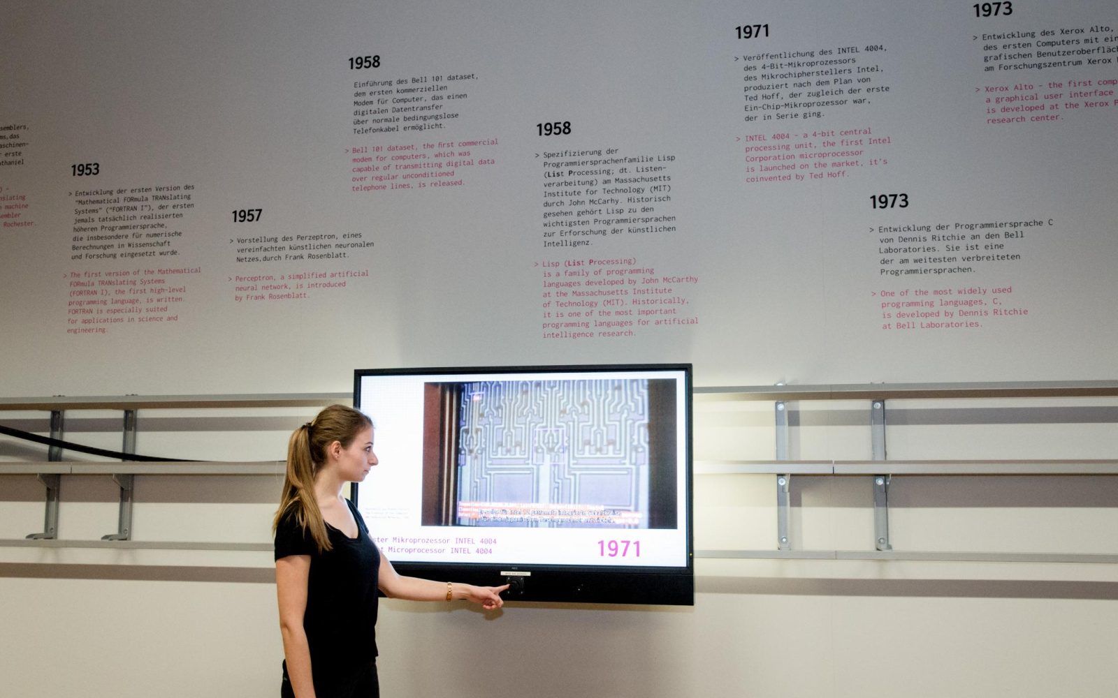 The picture shows a half-profile woman operating the virtual timeline of the »genalogy of the digital code«. On the wall, where the screen is mounted on a movable rail, you can see an information graphic