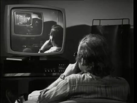 Zu sehen ist ein schwarz/weiß Bild, auf dem EIn älterer Mann sitzt vor einem Röhrenbildschirm auf dessen Bildschirm ein junger Mann zu sehen ist, der auch vor einem alten Fernsehr steht.