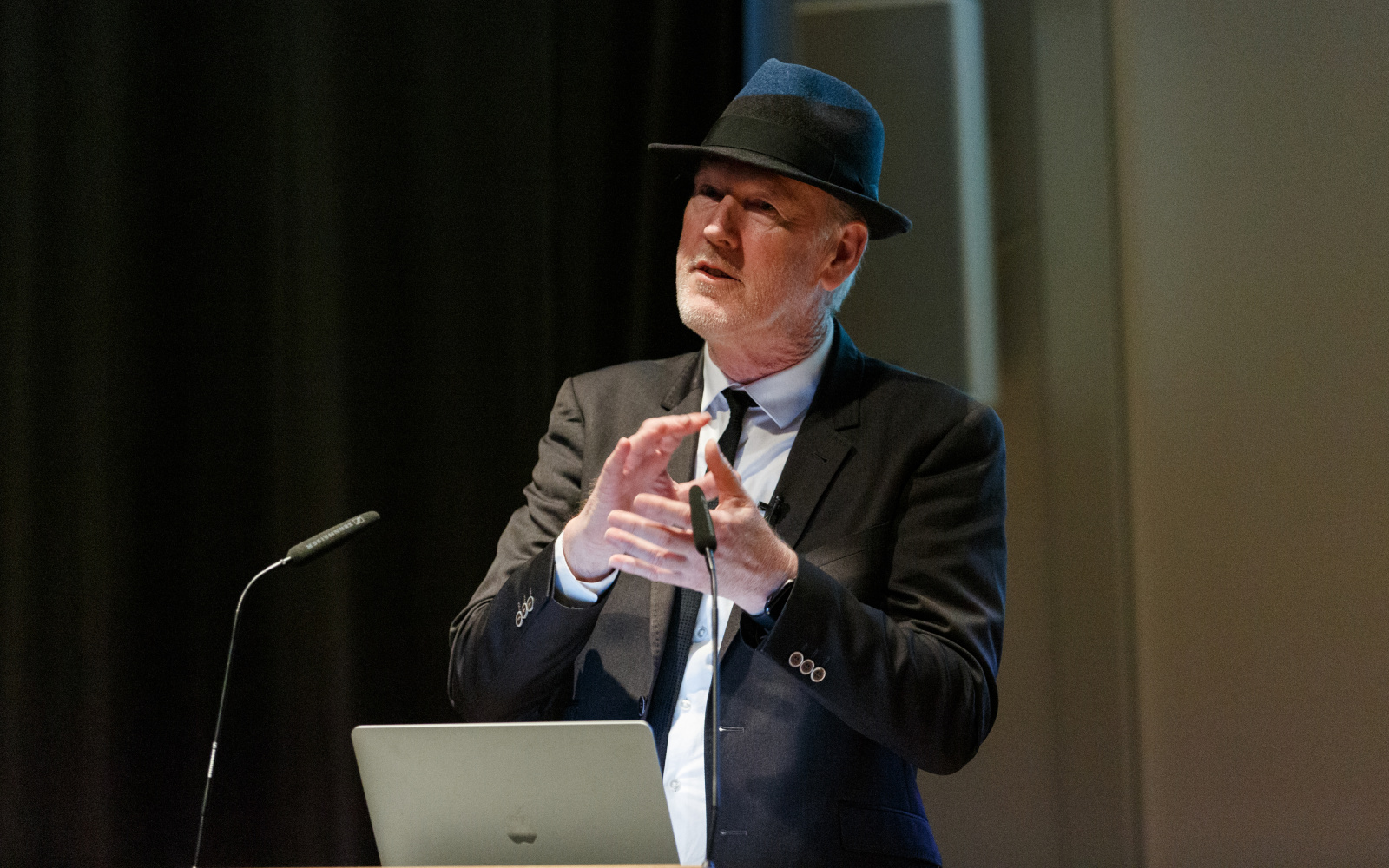 Zu sehen ist Thomas Paul, Medienkünstler und Professor in Anzug mit Hut, wie er hinter einem Rednerpult mit Laptop steht und gestikuliert.