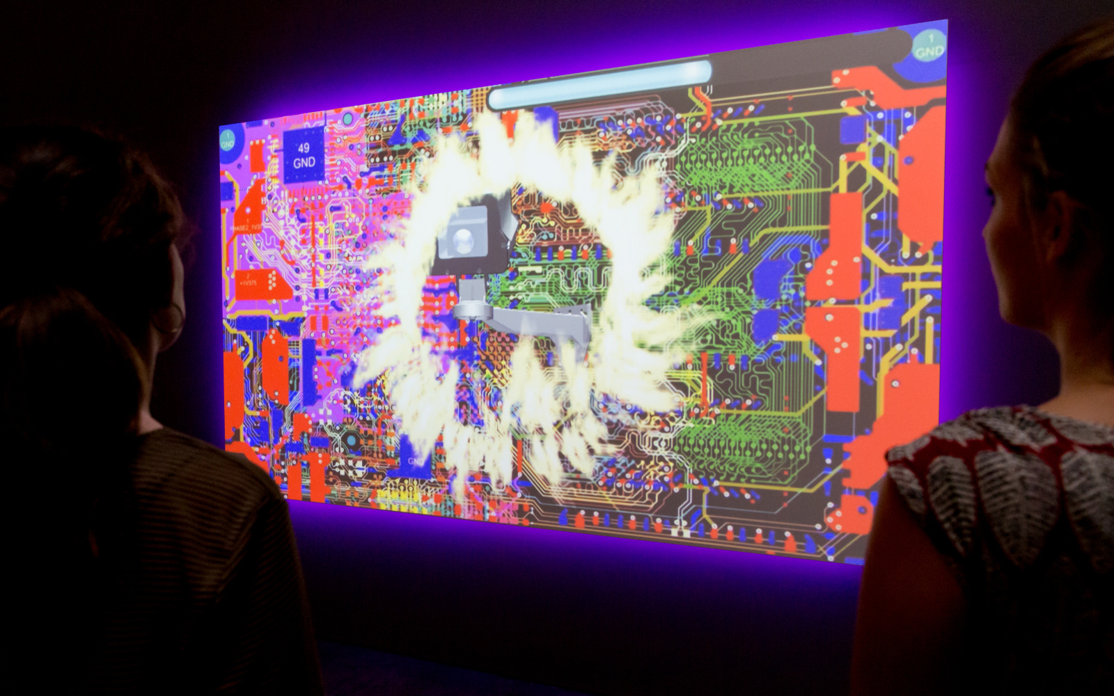Zwei Besucherinnen betrachten ein Video-Kunstwerk, das eine Collage aus Computer-Hardware und Flammen zeigt