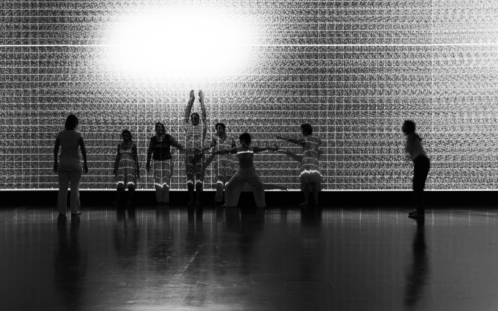 A group of dancers is moving in front of a wall with black and white projections.