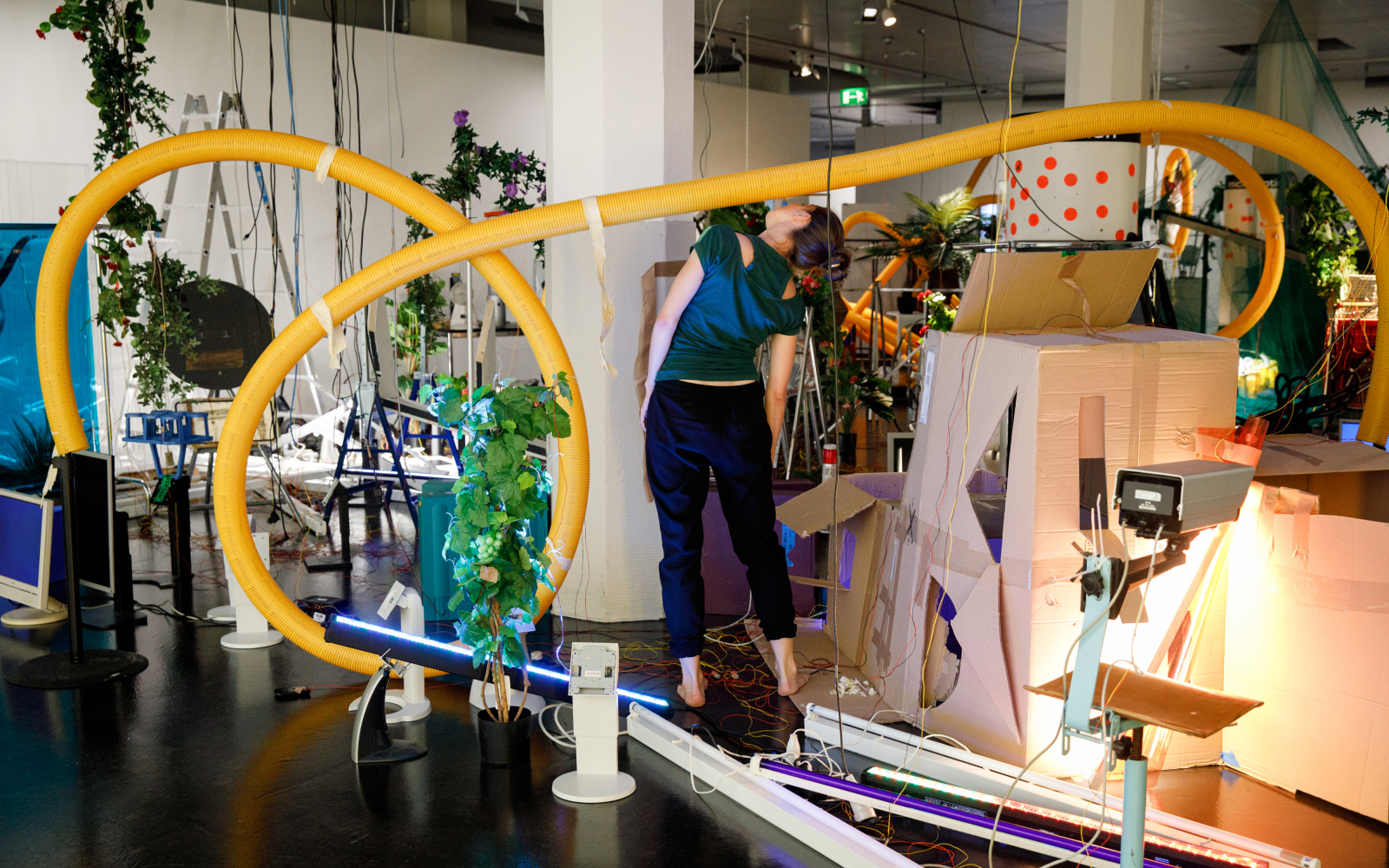 The photo shows an insight into the performance of the opening of »Edge of Now« in the midst of an artinalllation of plants, cardboard and plastic pipes.