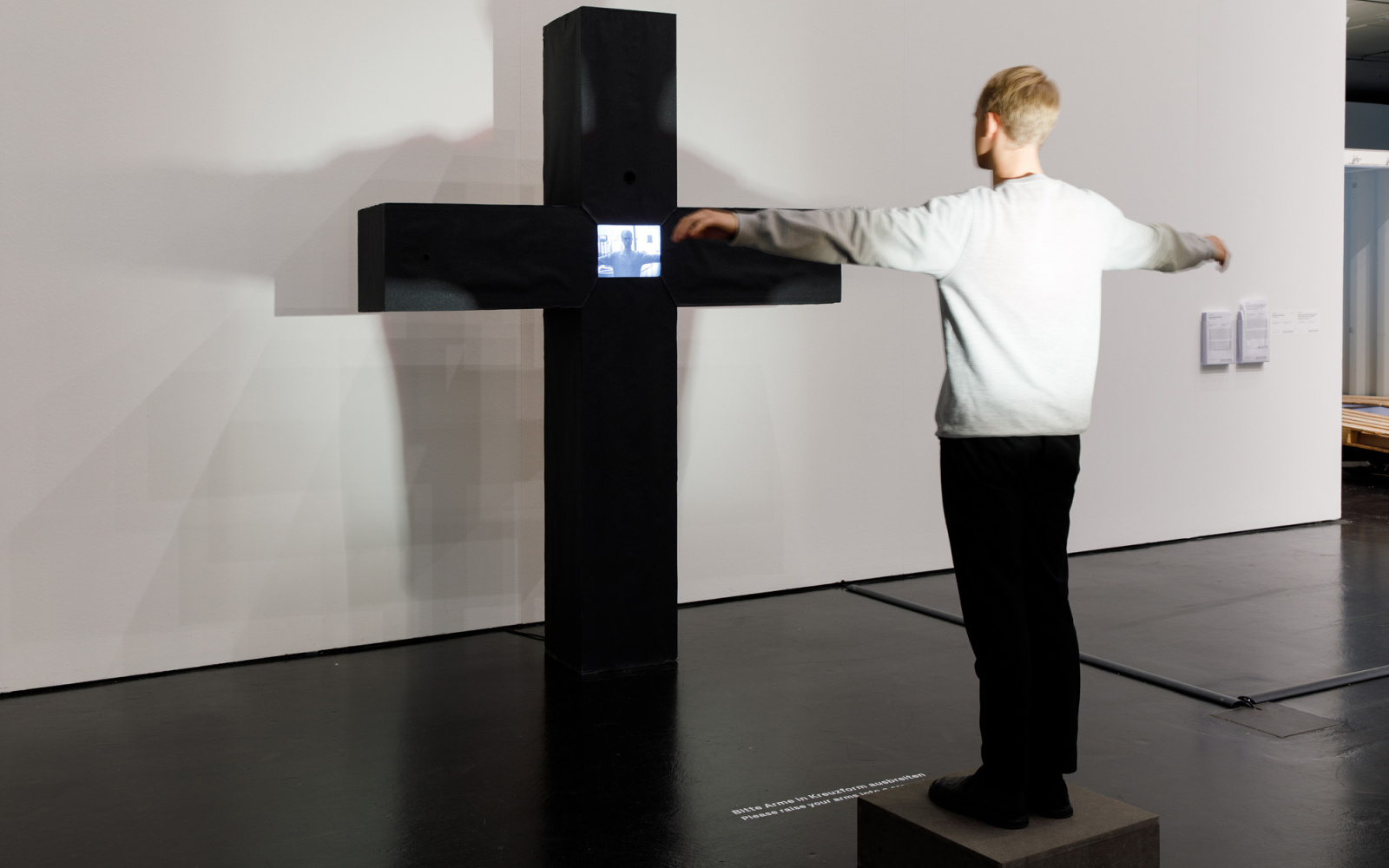 Peter Weibels interaktive Videoskultur: Ein schwarzes massives Kreuz. Dort wo sich die Streben des Kreuzes treffen, ist ein Monitor. Auf dem Monitor zu sehen ist die Person, die vor dem Kreuz steht und ihre Arme ausstreckt, als hinge sie selbst daran.