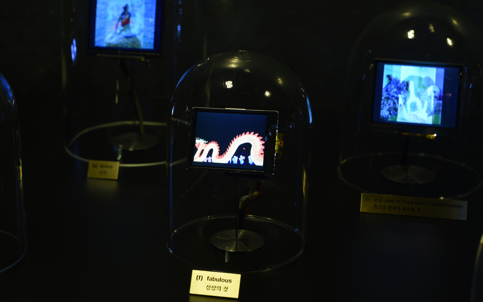 Das Foto zeigt eine Glasglocke in der ein kleiner Bildschirm steckt. Auf diesem ist ein Drachen ähnliches Wesen zu sehen.