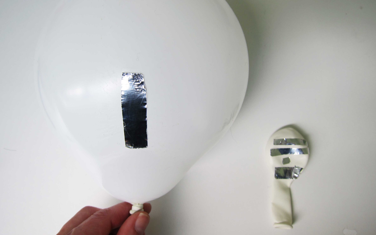 A white inflated balloon with a piece of shiny metallic tape on a white surface. Next to it a not inflated balloon with many pieces of tape, just like on the other one.